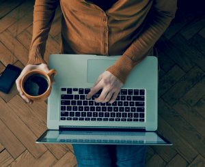 Woman sitting on the floor with a laptop & holding a cup of coffee.